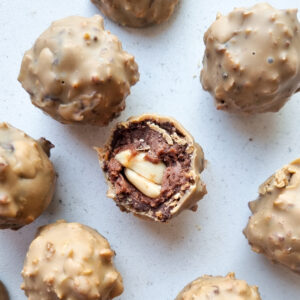 Pohľad zhora na pralinky príchute Peanut rocher a bočný prierez jednej pralinky. Obsah pralinky: Kakaová hmota obalená v bielej čokoláde a arašidovom masle, v strede pralinky je arašidový orech.