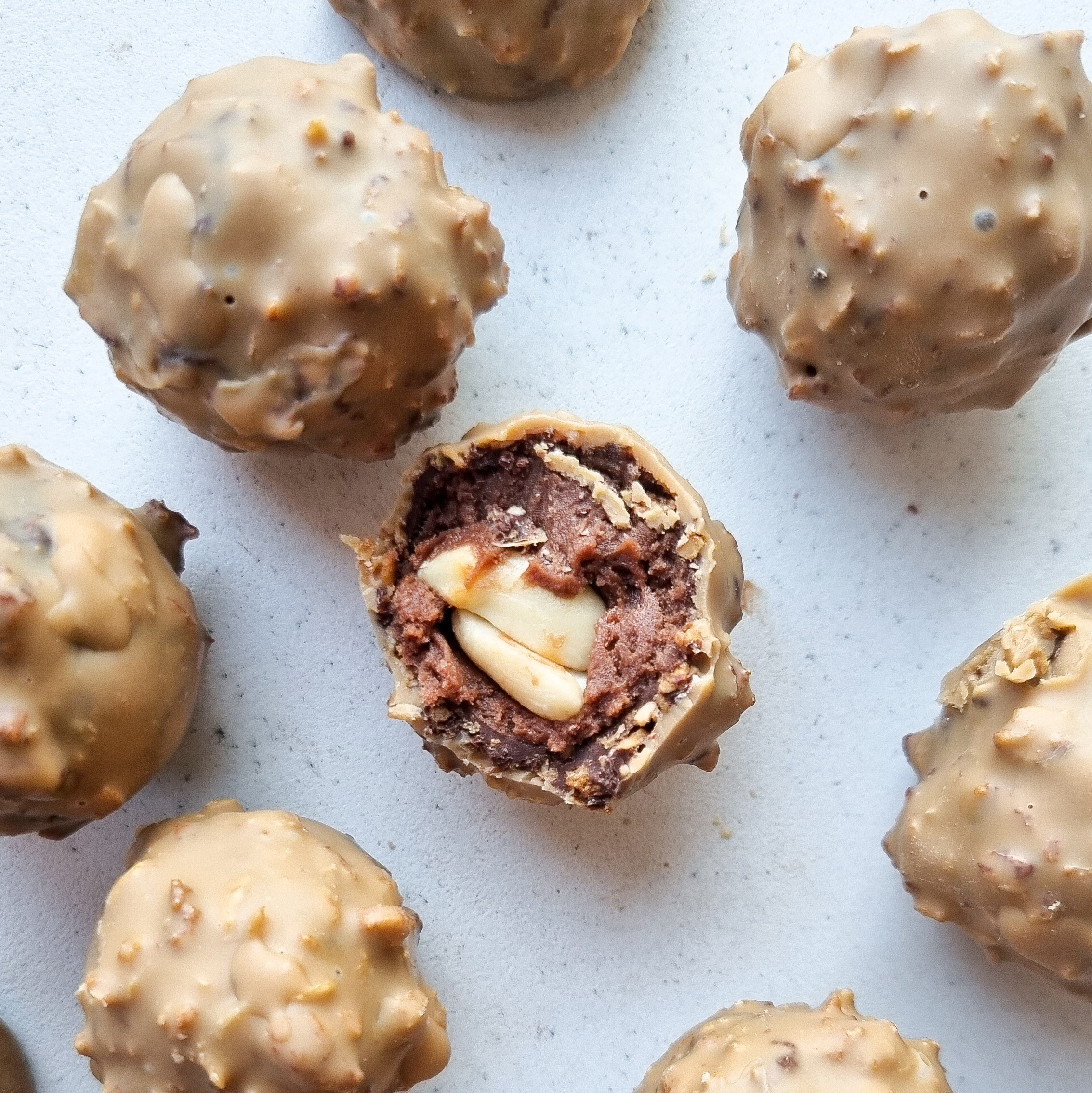 Pohľad zhora na pralinky príchute Peanut rocher a bočný prierez jednej pralinky. Obsah pralinky: Kakaová hmota obalená v bielej čokoláde a arašidovom masle, v strede pralinky je arašidový orech.